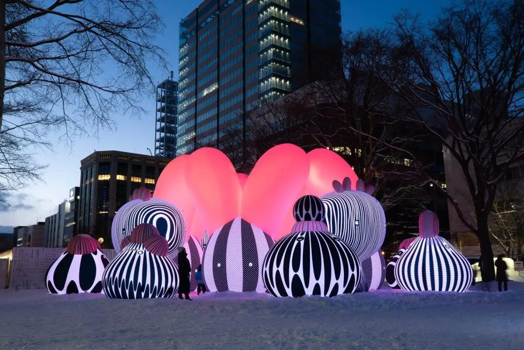 Colorful, illuminated inflatable sculptures create a vibrant outdoor art installation at dusk, featuring playful polka-dot patterns against a city skyline.