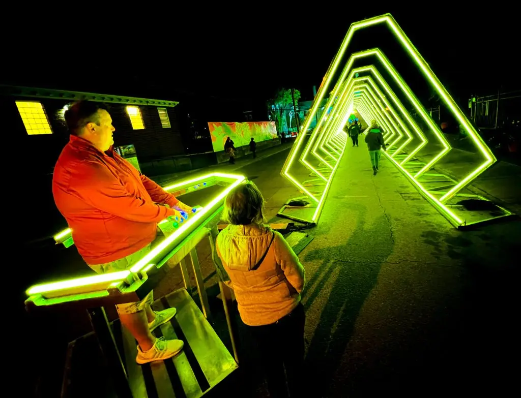 A person in a red jacket interacts with a glowing control panel beside a pathway lined with illuminated, neon green triangular arches. Another person stands nearby, looking at the arches. The scene is set in a nighttime setting with a vibrant, artistic ambiance.